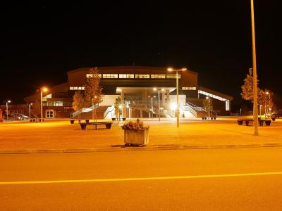 A large, modern sports hall illuminated at night, featuring a prominent central entrance with wide exterior staircases, a flat roofline with a row of lit windows, and a paved plaza in the foreground.