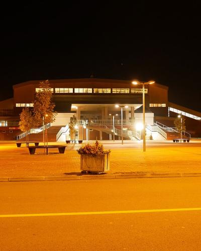 A large, modern sports hall illuminated at night, featuring a prominent central entrance with wide exterior staircases, a flat roofline with a row of lit windows, and a paved plaza in the foreground.