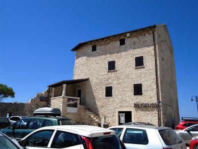 A tall, rectangular medieval stone tower with small shuttered windows, an attached lower structure with a staircase, and parked cars in the foreground under a clear blue sky.