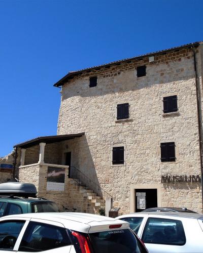 A tall, rectangular medieval stone tower with small shuttered windows, an attached lower structure with a staircase, and parked cars in the foreground under a clear blue sky.