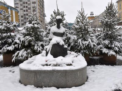 A snow-covered bronze statue of a seated woman on a circular stone pedestal, surrounded by snow-dusted evergreen trees in a paved urban square.