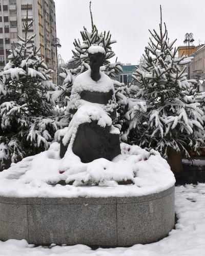 A snow-covered bronze statue of a seated woman on a circular stone pedestal, surrounded by snow-dusted evergreen trees in a paved urban square.
