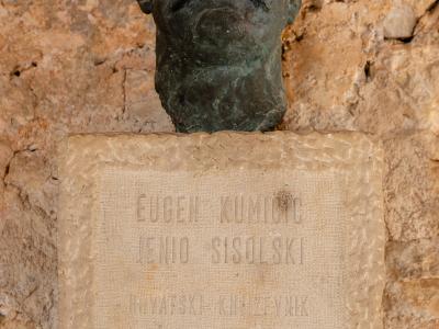 A bronze bust of a man with a mustache mounted on a stone plaque with engraved text, set against a rough stone wall.