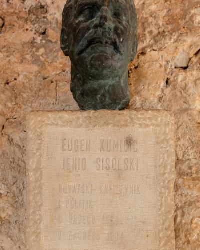 A bronze bust of a man with a mustache mounted on a stone plaque with engraved text, set against a rough stone wall.