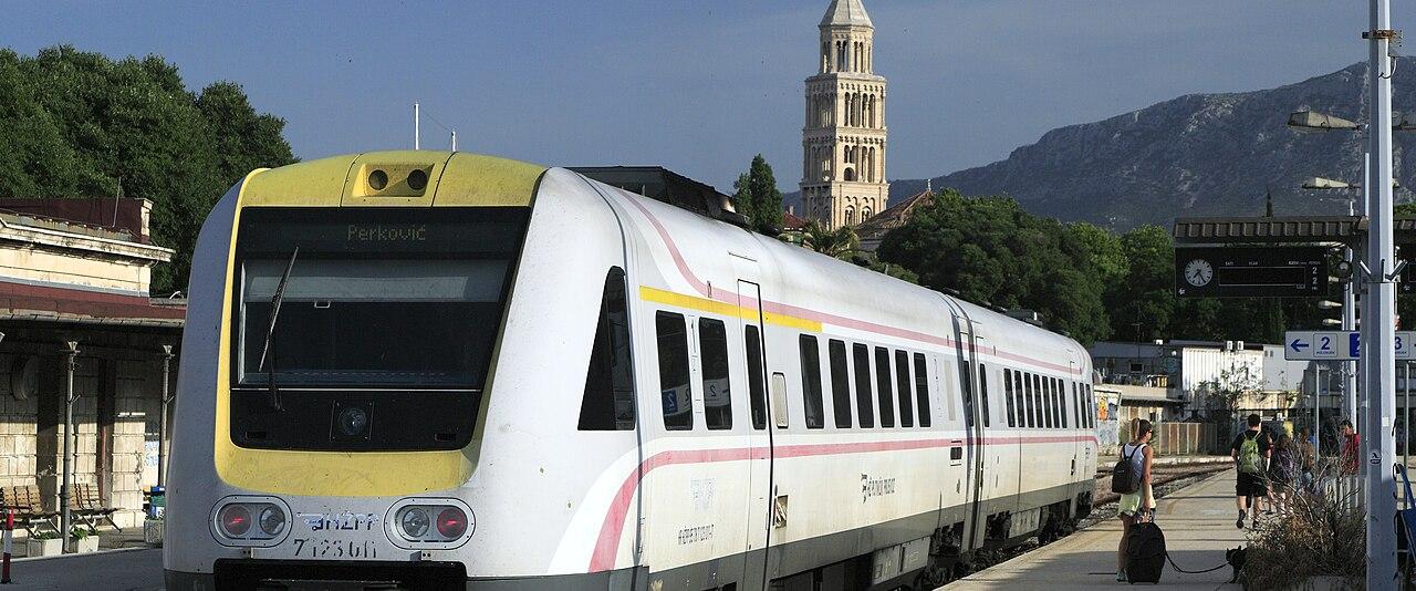 A white HŽPP diesel train at Split station with the cathedral bell tower behind
