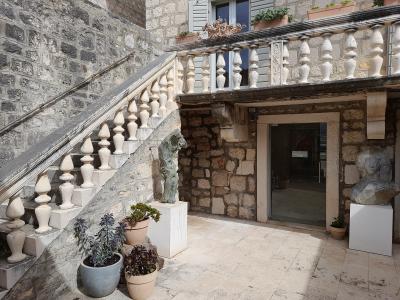 A stone courtyard featuring a staircase with a balustrade, potted plants, and two sculptures on white pedestals near an entrance doorway.