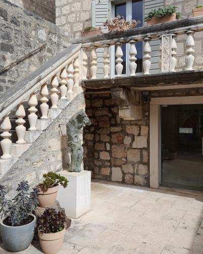 A stone courtyard featuring a staircase with a balustrade, potted plants, and two sculptures on white pedestals near an entrance doorway.