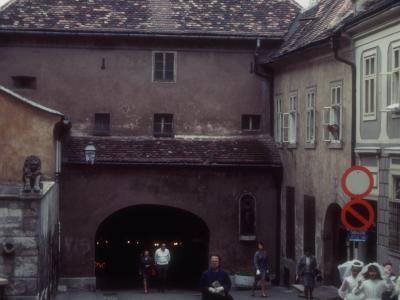 A medieval stone gate with a steep tiled roof, an arched passageway, and a statue niche on the right side, set in a narrow European street with pedestrians and two spires visible in the background.