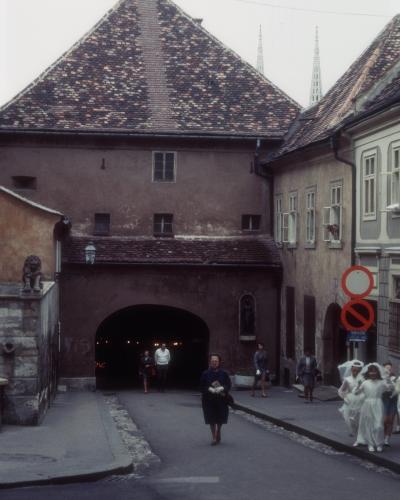 A medieval stone gate with a steep tiled roof, an arched passageway, and a statue niche on the right side, set in a narrow European street with pedestrians and two spires visible in the background.