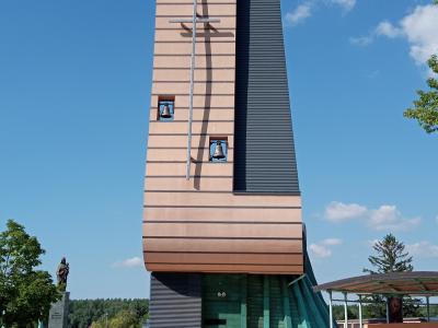 A modern church tower with a tall, rectangular beige facade featuring horizontal lines, three visible bells in square openings, a large metal cross mounted on the front, and a dark vertical panel on the right side; a statue stands to the left on a