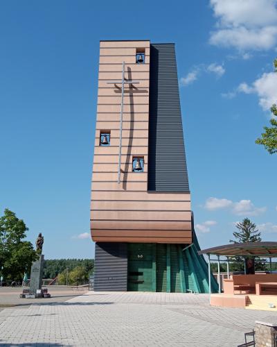 A modern church tower with a tall, rectangular beige facade featuring horizontal lines, three visible bells in square openings, a large metal cross mounted on the front, and a dark vertical panel on the right side; a statue stands to the left on a
