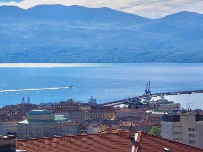 A panoramic view of the city of Rijeka, Croatia, showing the urban skyline with terracotta roofs, the Kvarner Bay, and the long Molo Lungo breakwater extending into the sea with cranes at its end, backed by the Učka mountain range.