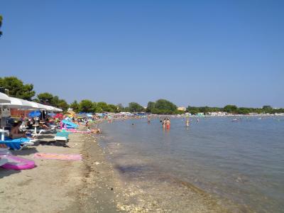 A crowded sandy beach with sunbathers, umbrellas, and people swimming in calm, clear water under a blue sky, with trees and buildings visible in the background.