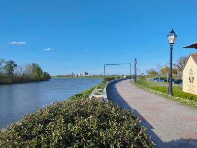 A paved riverside promenade with lamp posts and manicured hedges runs alongside a wide river, with a yellow building featuring a portrait and text on the right and industrial structures visible across the water.