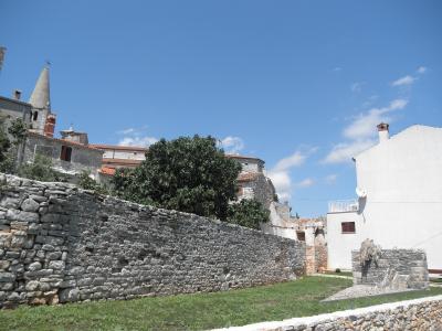 A low stone wall runs across the foreground with a grassy area, behind which are traditional Istrian stone buildings and a church tower; in the lower right corner is a small stone sculpture on a pedestal.