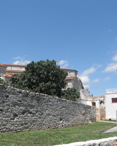 A low stone wall runs across the foreground with a grassy area, behind which are traditional Istrian stone buildings and a church tower; in the lower right corner is a small stone sculpture on a pedestal.