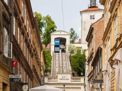 A narrow cobblestone street in Zagreb, Croatia, leading up to the Zagreb Funicular (labeled "ZET USPINJAČA") with its blue car ascending the track, flanked by historic buildings and outdoor café seating.