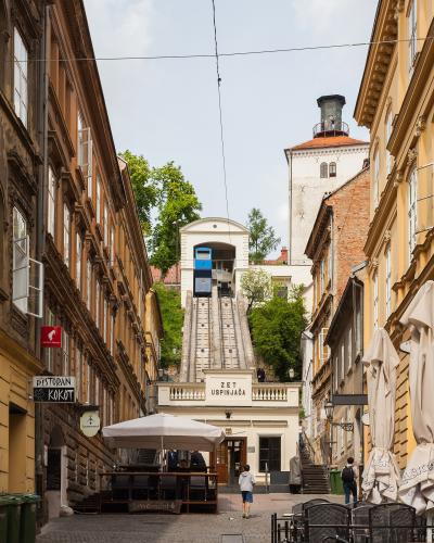 A narrow cobblestone street in Zagreb, Croatia, leading up to the Zagreb Funicular (labeled "ZET USPINJAČA") with its blue car ascending the track, flanked by historic buildings and outdoor café seating.