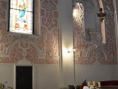 The image shows the interior of a Baroque-style church with ornate pink and white stucco decorations, a stained-glass window depicting a saint, a marble altar with candles, and a checkered floor.