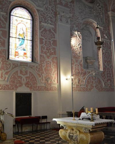 The image shows the interior of a Baroque-style church with ornate pink and white stucco decorations, a stained-glass window depicting a saint, a marble altar with candles, and a checkered floor.
