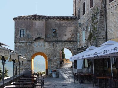 A stone archway gate with a yellow-painted interior arch, flanked by outdoor cafe seating under umbrellas, with a view of a valley through the arch.