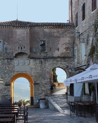 A stone archway gate with a yellow-painted interior arch, flanked by outdoor cafe seating under umbrellas, with a view of a valley through the arch.