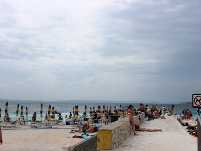A beach scene with people sunbathing on a paved promenade and sandy area, featuring rows of blue and yellow striped umbrellas and lounge chairs under a cloudy sky.