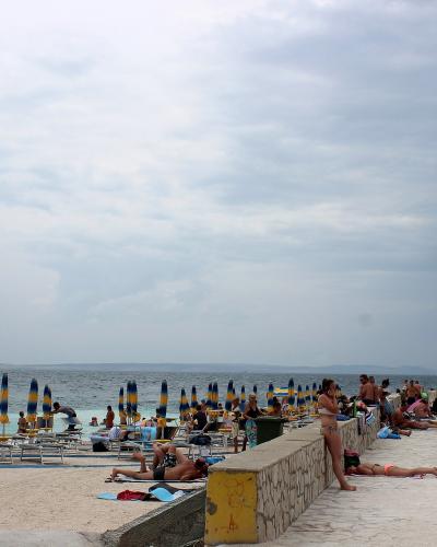 A beach scene with people sunbathing on a paved promenade and sandy area, featuring rows of blue and yellow striped umbrellas and lounge chairs under a cloudy sky.