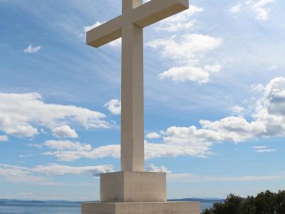 A large, modern white stone cross stands on a tiered stone base atop a hill, overlooking the sea and distant islands under a blue sky with scattered clouds.