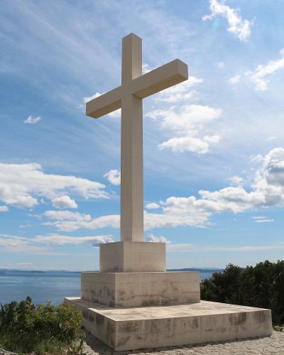 A large, modern white stone cross stands on a tiered stone base atop a hill, overlooking the sea and distant islands under a blue sky with scattered clouds.