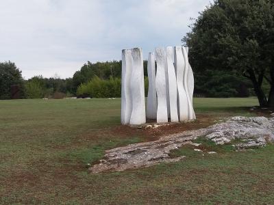 A group of tall, white, abstract stone sculptures standing on a grassy lawn with trees and other sculptures in the background.