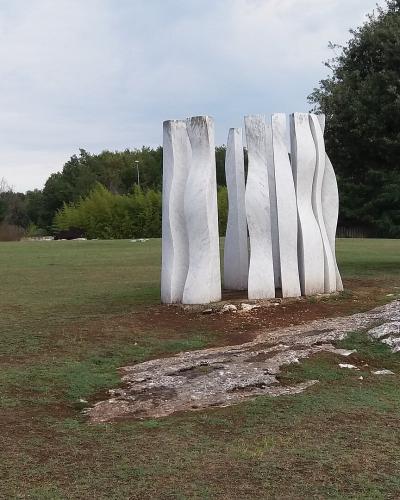 A group of tall, white, abstract stone sculptures standing on a grassy lawn with trees and other sculptures in the background.