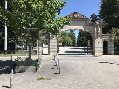 A glass sign reading "PARK MAKSIMIR" stands in the foreground next to a row of bollards, with the ornate stone entrance gate of the park visible in the background under a clear blue sky.