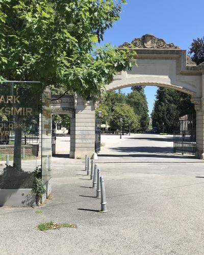 A glass sign reading "PARK MAKSIMIR" stands in the foreground next to a row of bollards, with the ornate stone entrance gate of the park visible in the background under a clear blue sky.