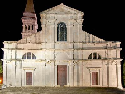 A night photograph of the illuminated Baroque facade of a large stone cathedral with a prominent bell tower to the left, featuring three doors and a large central arched window.
