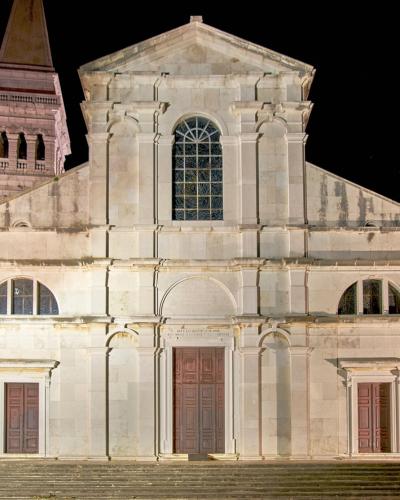 A night photograph of the illuminated Baroque facade of a large stone cathedral with a prominent bell tower to the left, featuring three doors and a large central arched window.