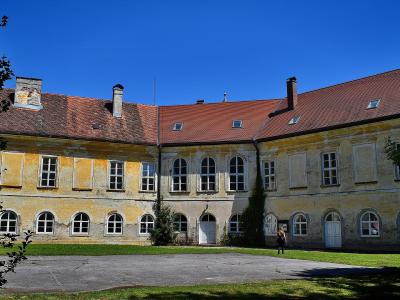 A yellow Baroque-style building with a red tiled roof, white-framed windows, and a paved courtyard in the foreground under a clear blue sky.