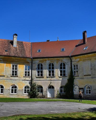 A yellow Baroque-style building with a red tiled roof, white-framed windows, and a paved courtyard in the foreground under a clear blue sky.