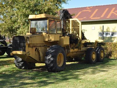 A large yellow forestry harvester machine parked on a grassy lawn in front of a building with a red roof.