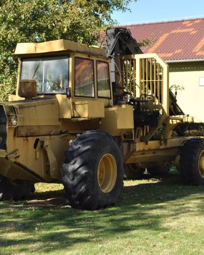 A large yellow forestry harvester machine parked on a grassy lawn in front of a building with a red roof.