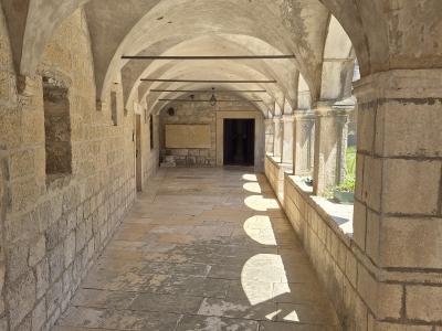 A stone cloister with vaulted ceilings and arched openings, featuring a paved walkway and sunlight casting circular shadows on the floor.