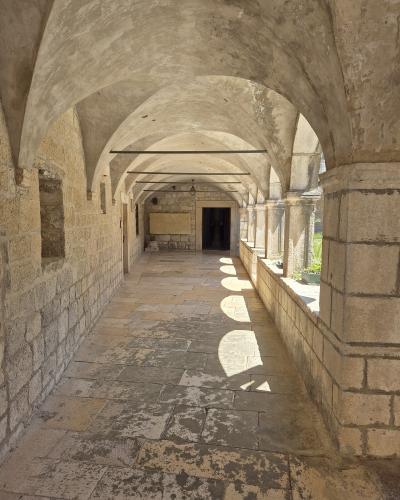 A stone cloister with vaulted ceilings and arched openings, featuring a paved walkway and sunlight casting circular shadows on the floor.