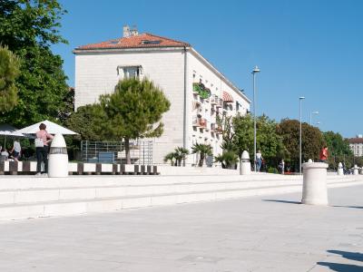 A wide, paved waterfront promenade with tiered white stone seating, modern bollards, and a large white building with a red-tiled roof in the background under a clear blue sky.