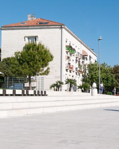 A wide, paved waterfront promenade with tiered white stone seating, modern bollards, and a large white building with a red-tiled roof in the background under a clear blue sky.