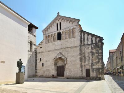 A close-up architectural view of the stone apse of a Romanesque church, featuring a blind arcade of columns and arches under a conical tiled roof against a blue sky.