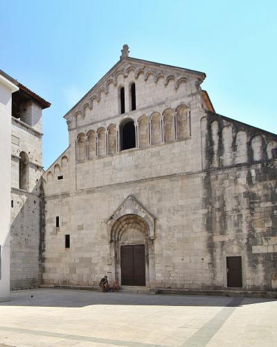 A close-up architectural view of the stone apse of a Romanesque church, featuring a blind arcade of columns and arches under a conical tiled roof against a blue sky.