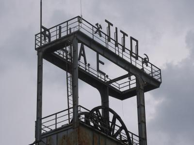 A rusted steel headframe of an old mine shaft, featuring large metal letters spelling "TITO" and "RAE" (likely part of "RAJE" or "RAJEVAC") mounted on the upper platform, with a hammer and sickle symbol visible near the top right.