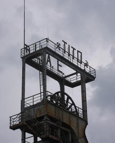 A rusted steel headframe of an old mine shaft, featuring large metal letters spelling "TITO" and "RAE" (likely part of "RAJE" or "RAJEVAC") mounted on the upper platform, with a hammer and sickle symbol visible near the top right.