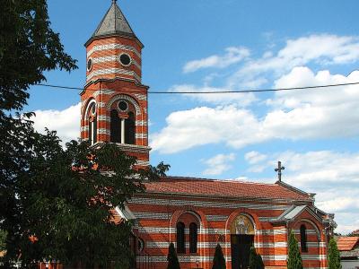 A red brick Orthodox church with white horizontal stripes, a tall bell tower topped with a cross, and a smaller cross on the main roof, set against a blue sky with clouds.