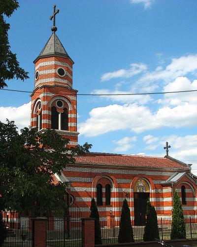 A red brick Orthodox church with white horizontal stripes, a tall bell tower topped with a cross, and a smaller cross on the main roof, set against a blue sky with clouds.
